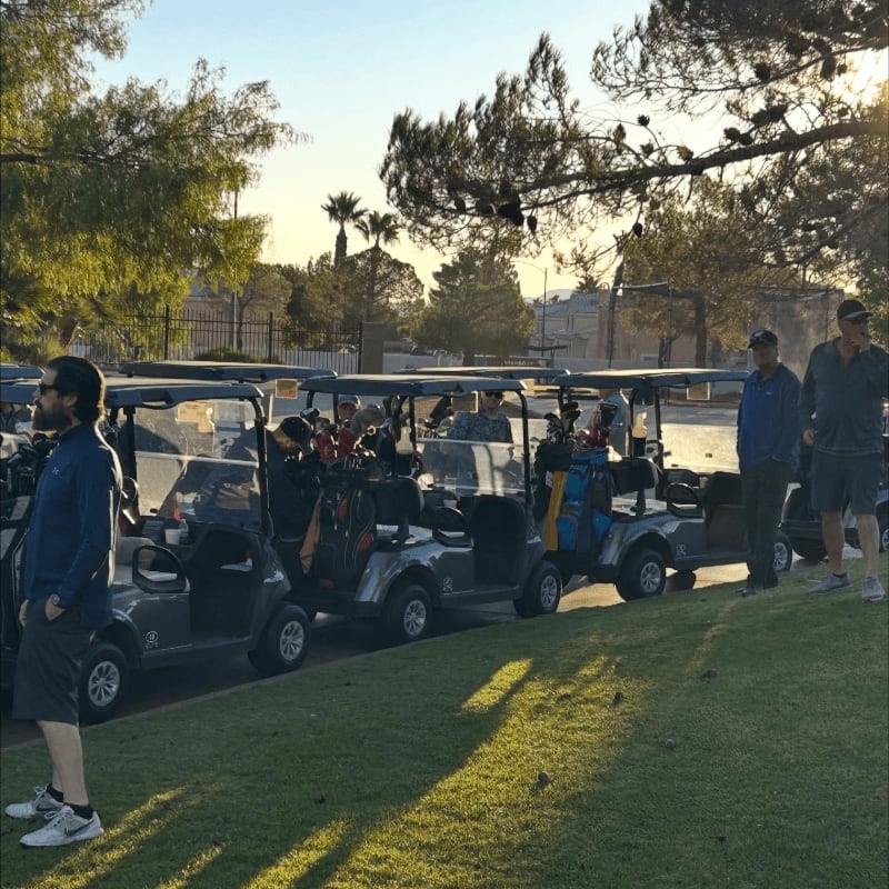 A man stands in the foreground on a grassy area next to a line of parked golf carts, with trees and buildings visible in the sunlit background.