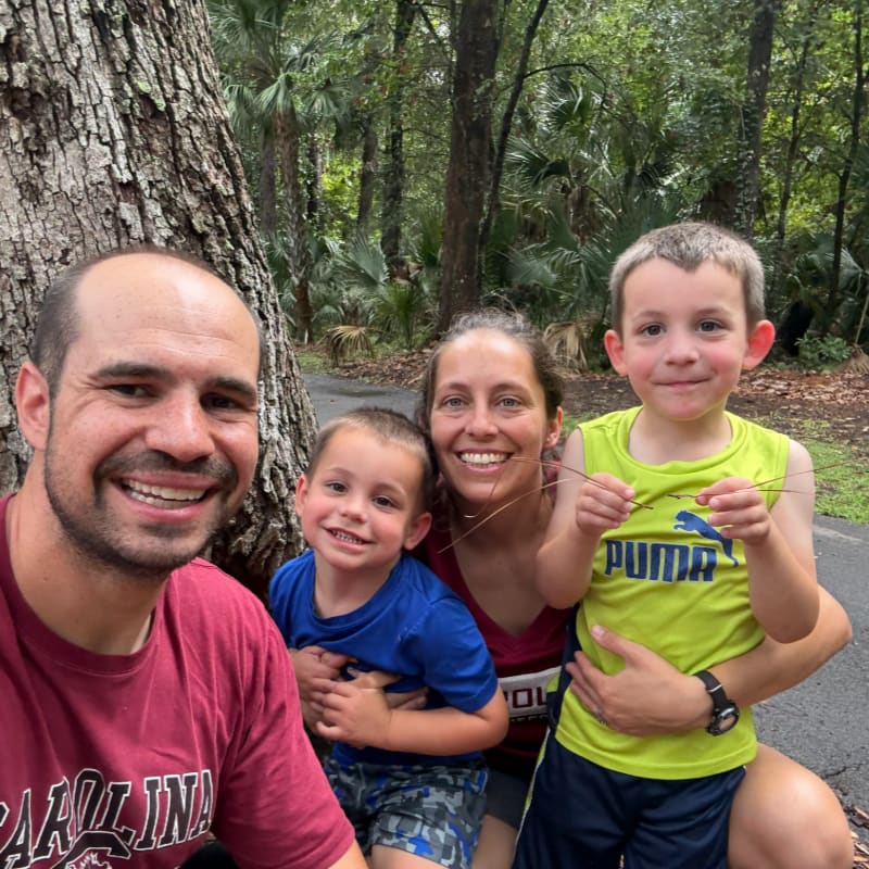 A smiling family of four poses for a selfie in front of a large tree, with a lush, green forest in the background.