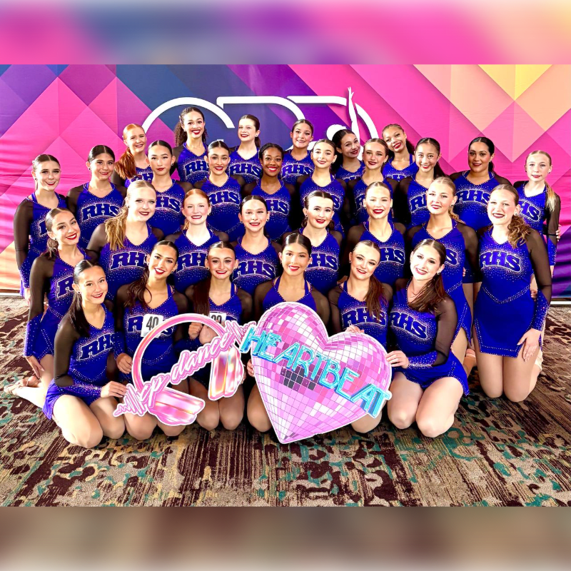 A dance team in matching blue uniforms poses in front of a geometric, pink and purple backdrop, holding a large pink heart prop.