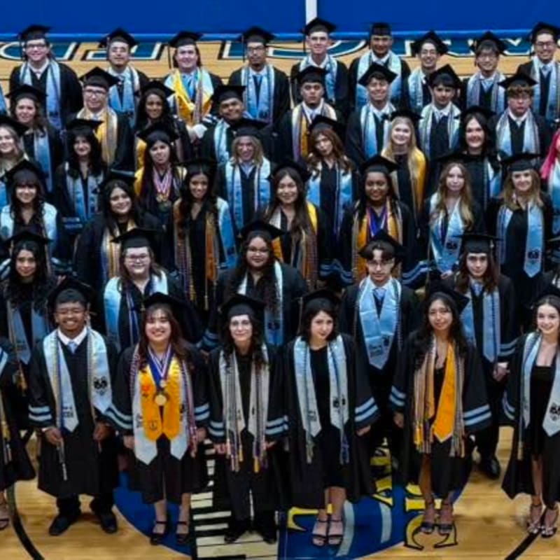 A large group of smiling graduates in black gowns and caps pose for a photo on a blue and yellow gymnasium floor, with a blue banner hanging in the background.