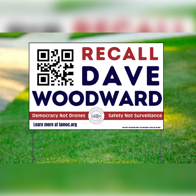 A yard sign with a QR code and the words "RECALL DAVE WOODWARD" is planted in green grass, with a blurred background of a sidewalk and trees.