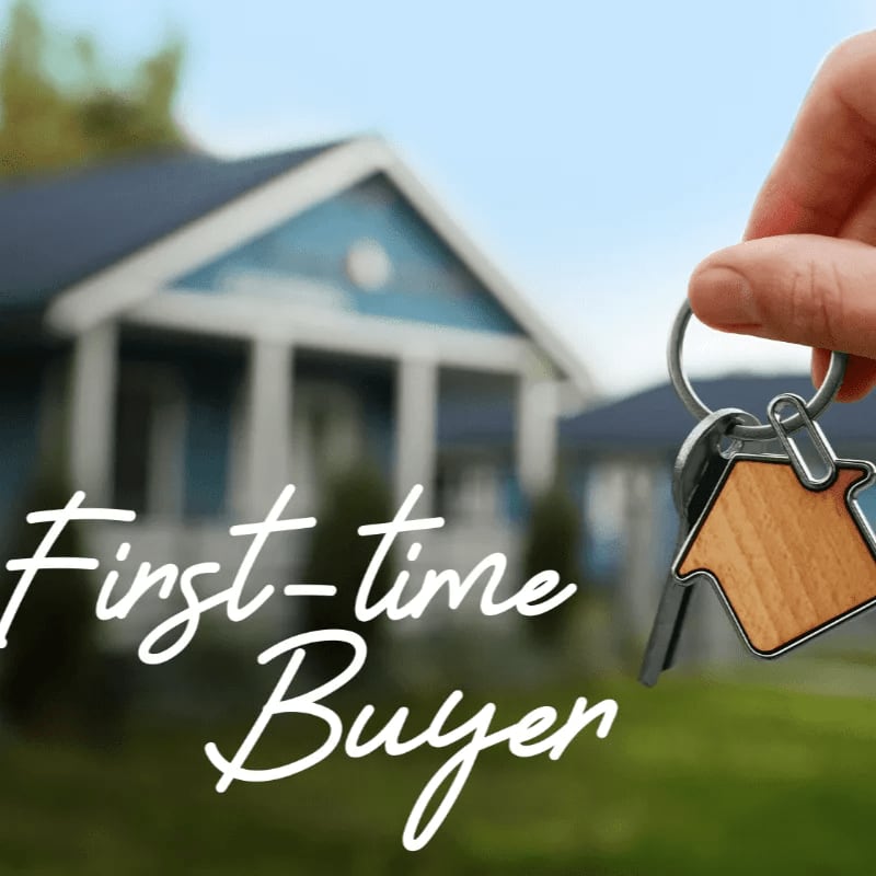 A hand holds a house-shaped keychain with keys in front of a blurred blue house under a bright sky.