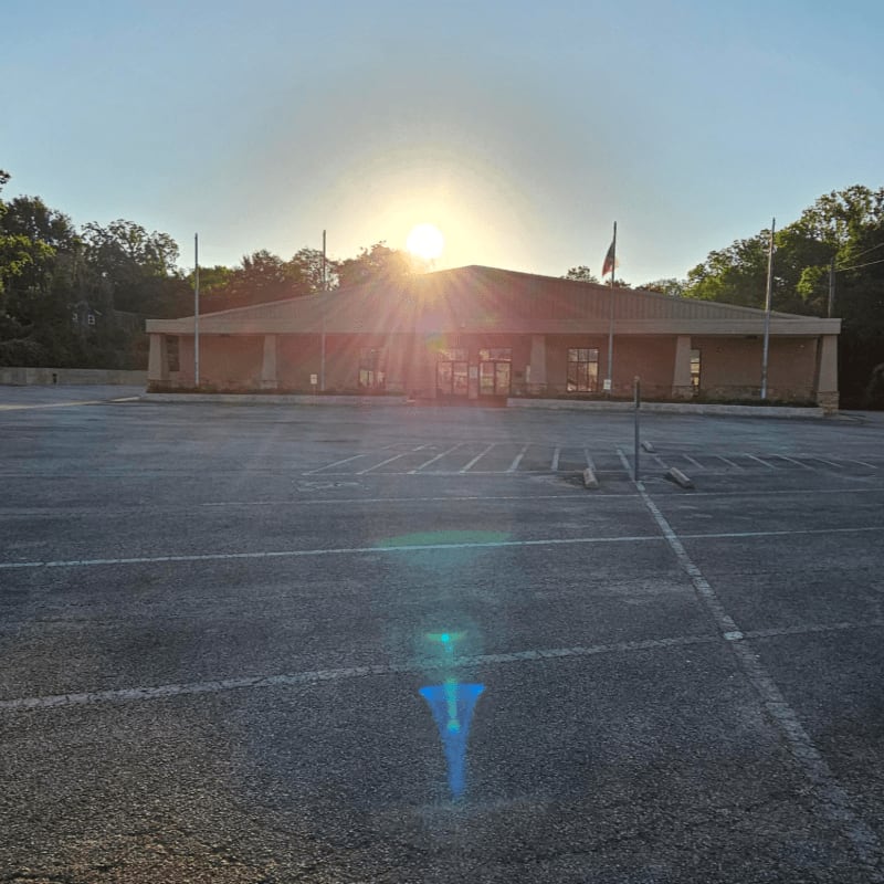 An empty parking lot stretches towards a sun-drenched building framed by trees.
