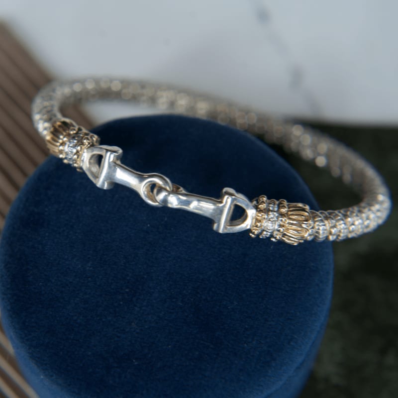 A silver and gold bracelet with a bit clasp rests on a dark blue velvet surface, with a blurred background of textured fabric.