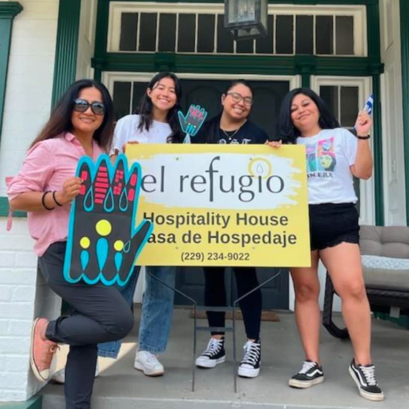 Four smiling women pose with a yellow sign that reads "El Refugio Hospitality House" in front of a white and green house.