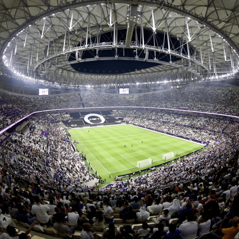 A packed stadium with a vibrant green soccer field in the center, surrounded by a sea of spectators under a modern, illuminated roof.