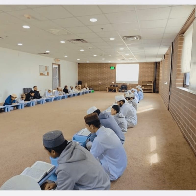 In the foreground, young boys in kufis sit on the floor reading from books, while in the background, a group of men are seated in a classroom setting.