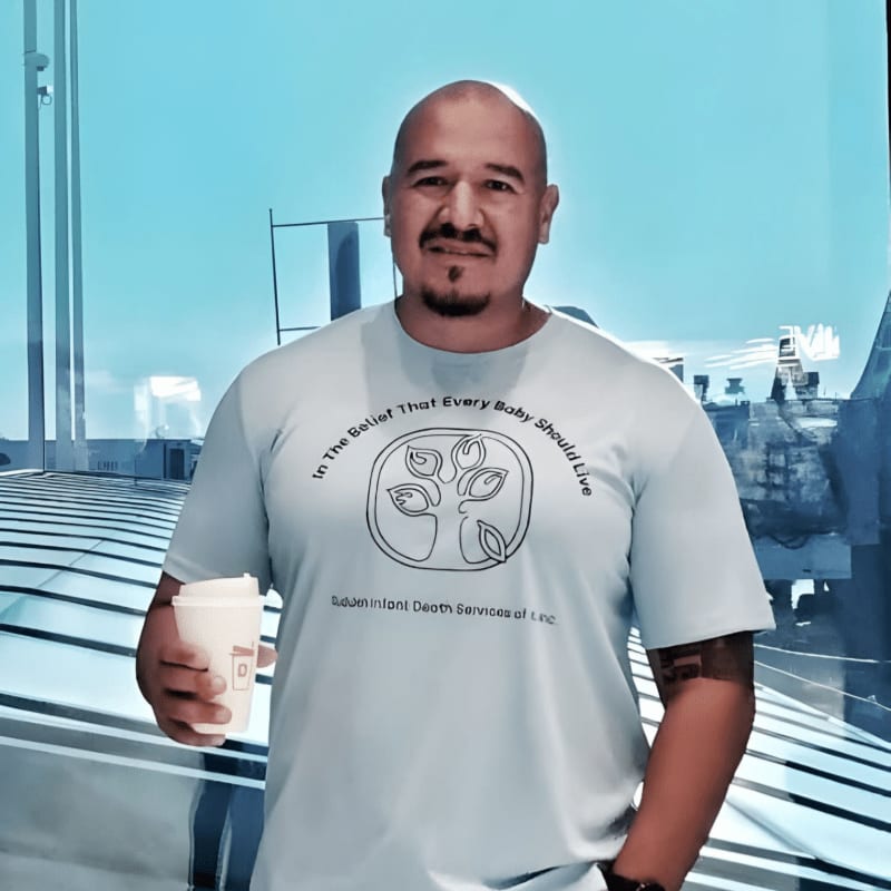 A bald man with a goatee and a light blue t-shirt stands holding a coffee cup in front of a large window with a city skyline in the background.