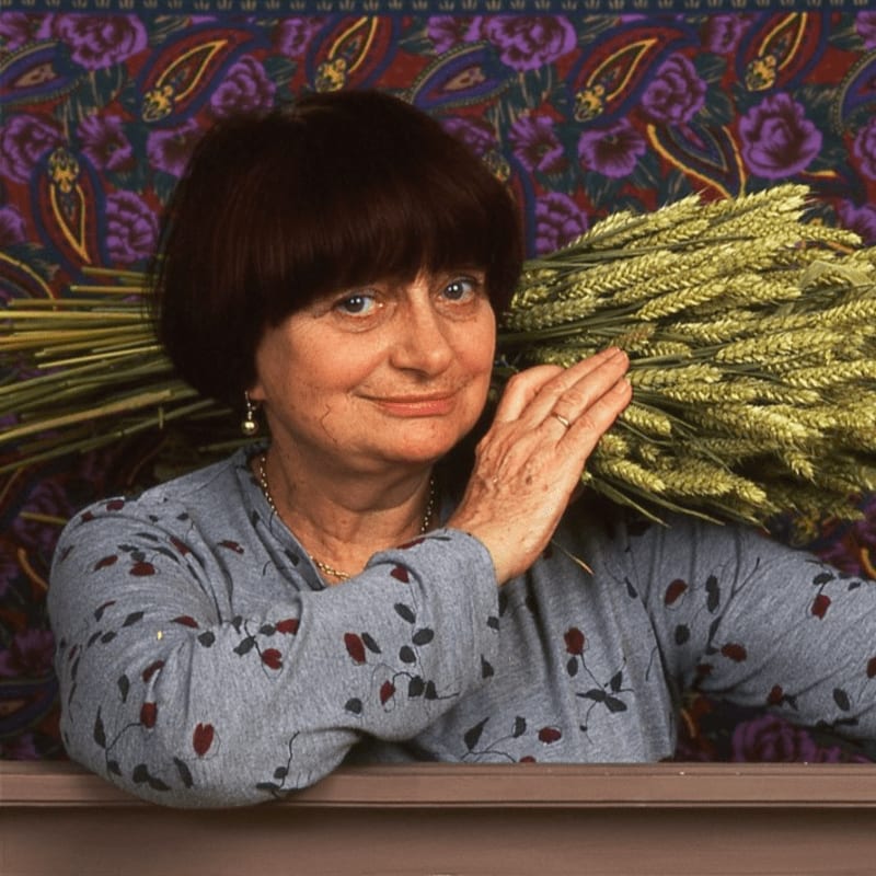A woman with a bowl cut and a patterned shirt carries a bundle of wheat over her shoulder against a vibrant paisley background.