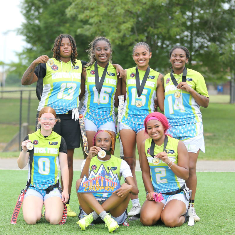 Six young women in athletic uniforms with medals stand and kneel on a grassy field with a blurred background of trees and a fence.