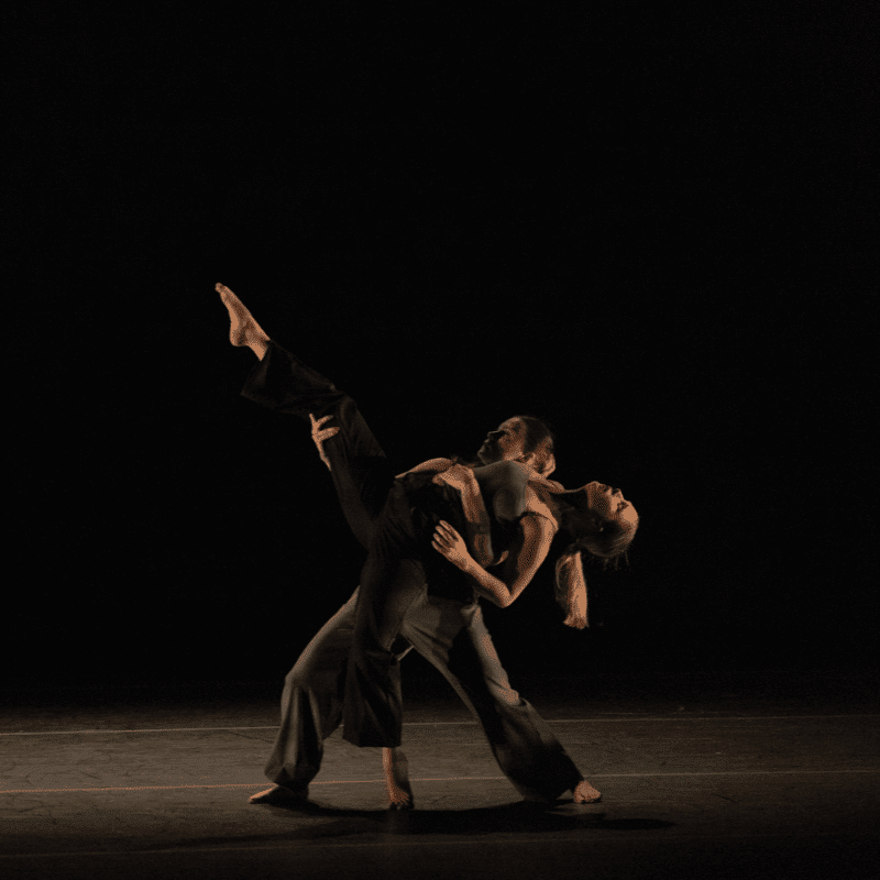 A male dancer supports a female dancer in a dramatic pose on a dark stage.