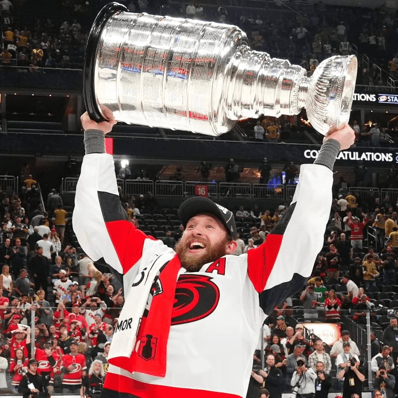 A jubilant hockey player in a white and red jersey hoists the Stanley Cup above his head in a crowded arena.