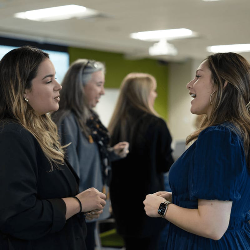 Two women in the foreground are engaged in conversation, while other individuals are blurred in the background of an office setting.