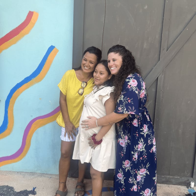 Three women pose for a photo in front of a colorful mural and a weathered wooden door.