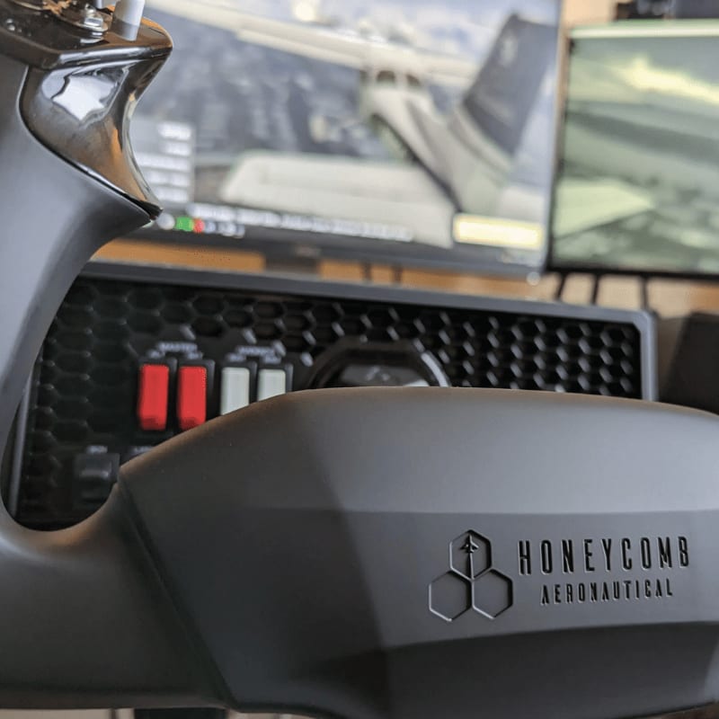A close-up of a grey flight simulator yoke with the "Honeycomb Aeronautical" logo in the foreground, and two computer monitors displaying an airplane cockpit in the background.