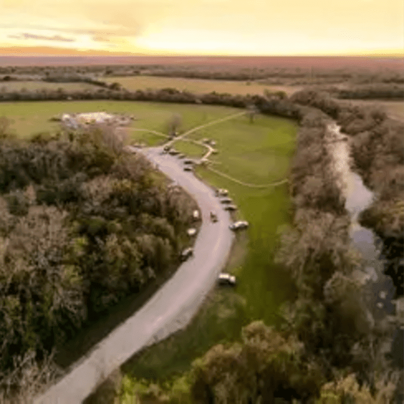 A winding road with parked cars leads through a grassy field bordered by trees, with a river on the right and a hazy sunset in the distance.