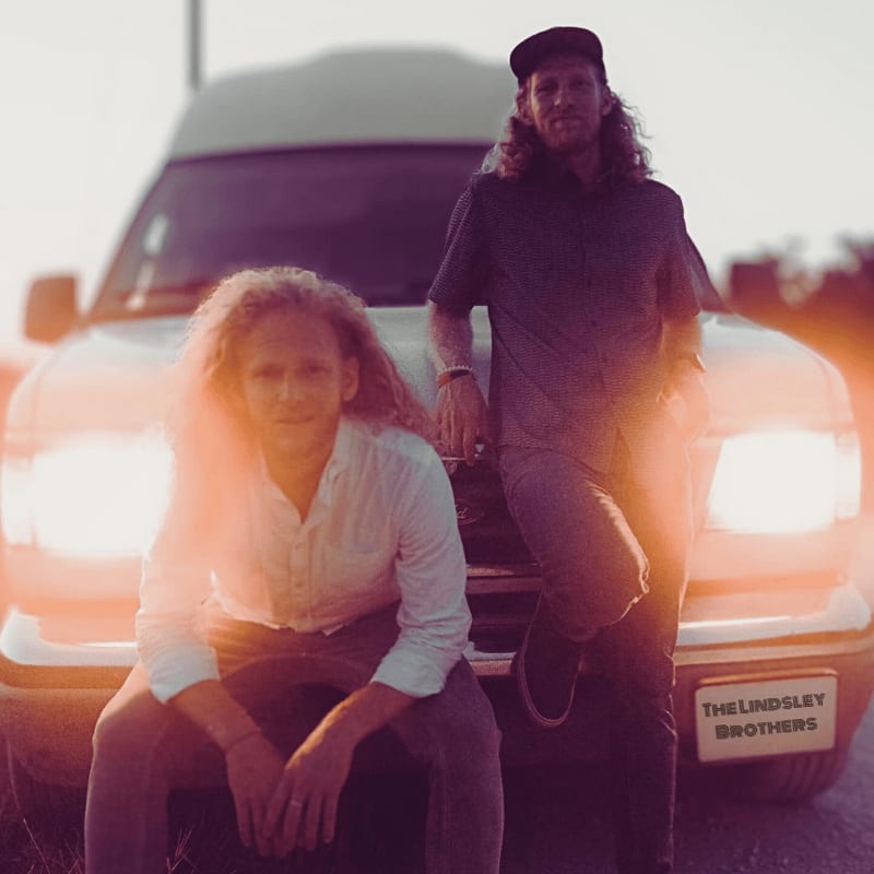 Two men with long, curly hair pose in front of a van with its headlights on, set against a hazy, sunlit background.