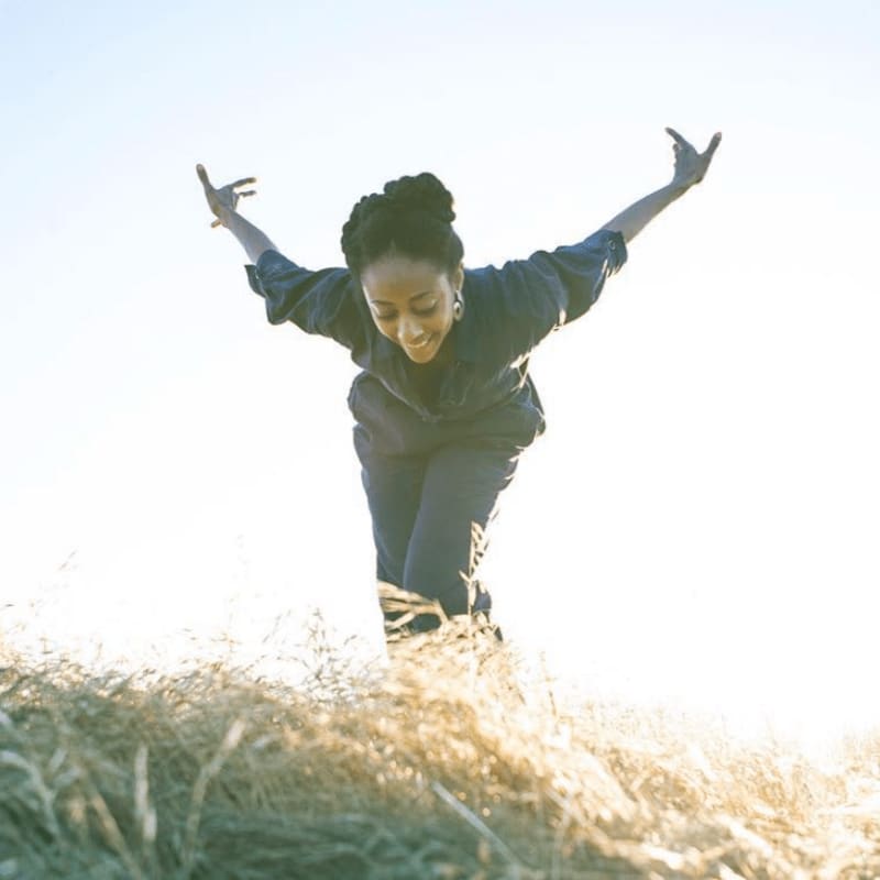 A woman with arms outstretched smiles as she stands in tall, golden grass against a bright, hazy sky.