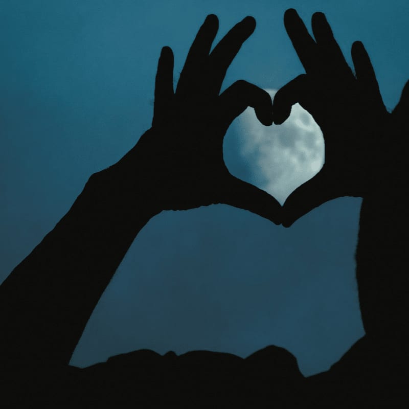 Silhouetted hands form a heart shape in the foreground, framing a full moon against a dark blue sky in the background.