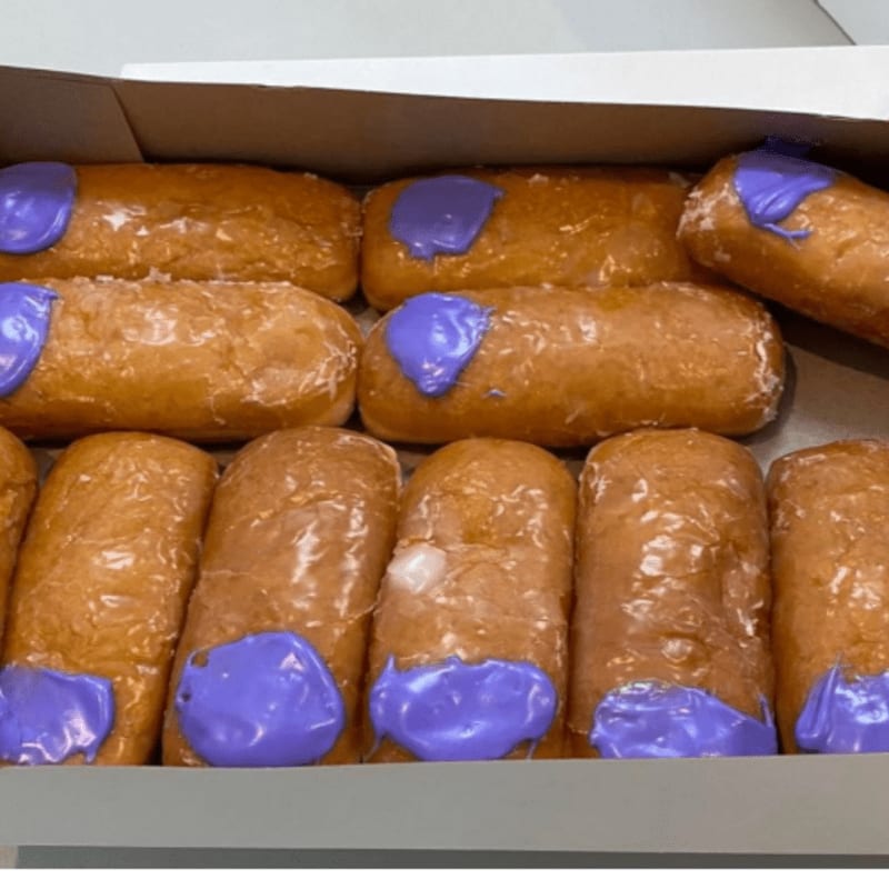 A box is filled with glazed donuts topped with purple icing in the foreground, with the white interior of the box visible in the background.