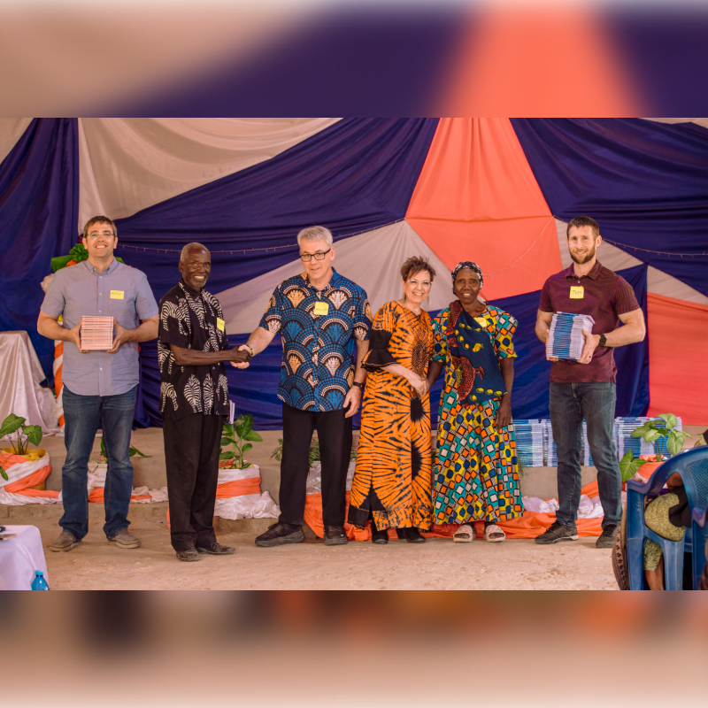 Six people stand in the foreground holding gifts, with a colorful draped fabric backdrop behind them.