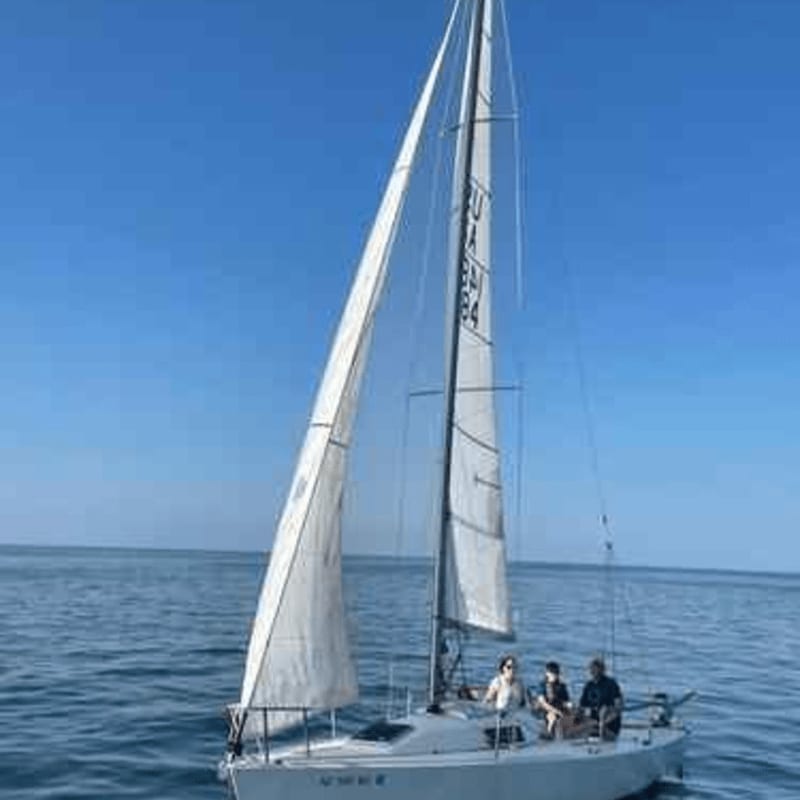 A white sailboat with three people aboard sails on calm blue water under a clear sky.