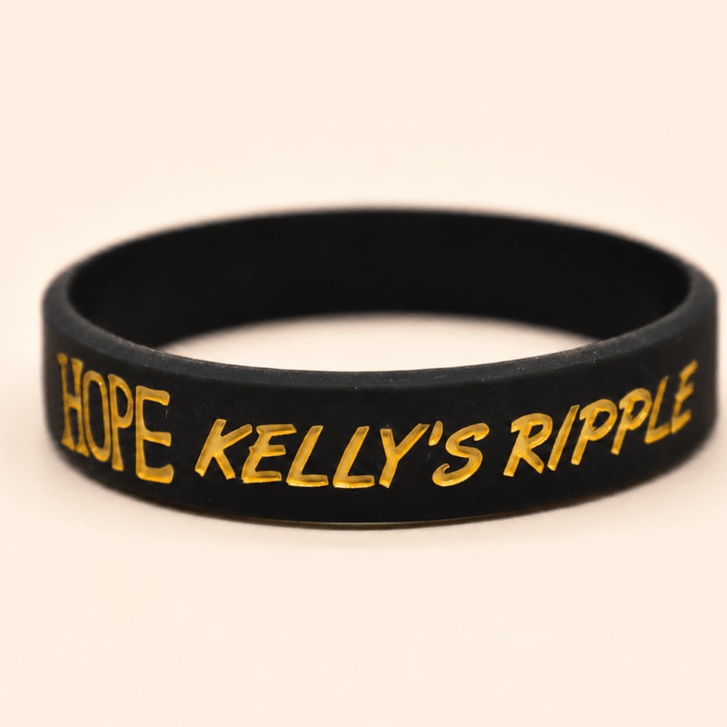 A black silicone wristband with gold lettering reading "HOPE KELLY'S RIPPLE" rests on a plain, light-colored surface.