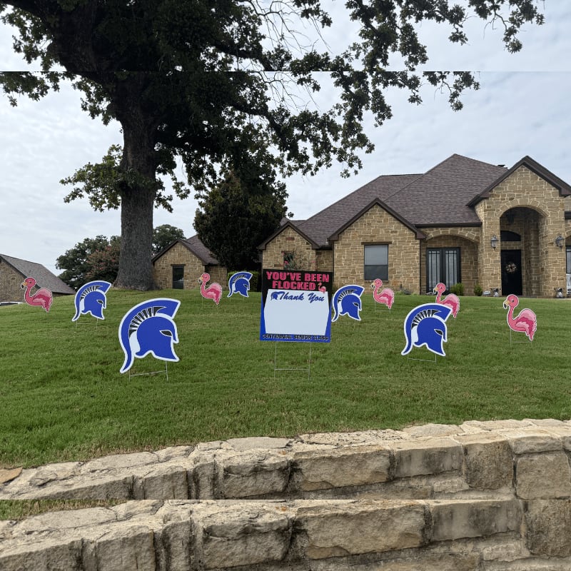 Pink flamingos and blue Spartan helmets dot a grassy lawn in front of a stone house, with a large tree and cloudy sky in the background.