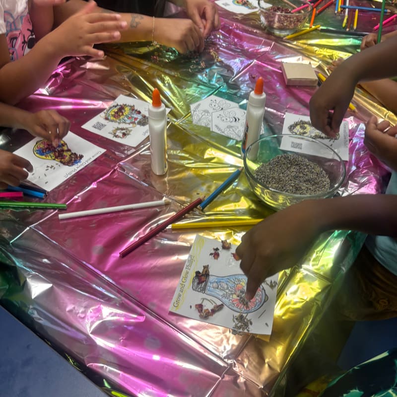 Children's hands are busy with craft supplies on a rainbow-colored tablecloth, with a bowl of seeds and coloring pages in the foreground and more craft materials in the background.