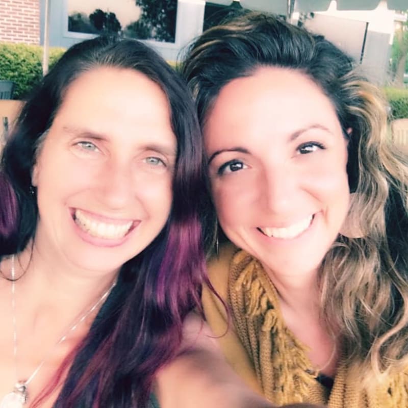 Two smiling women with purple-streaked hair in the foreground are captured against a blurred outdoor background with greenery and a building.