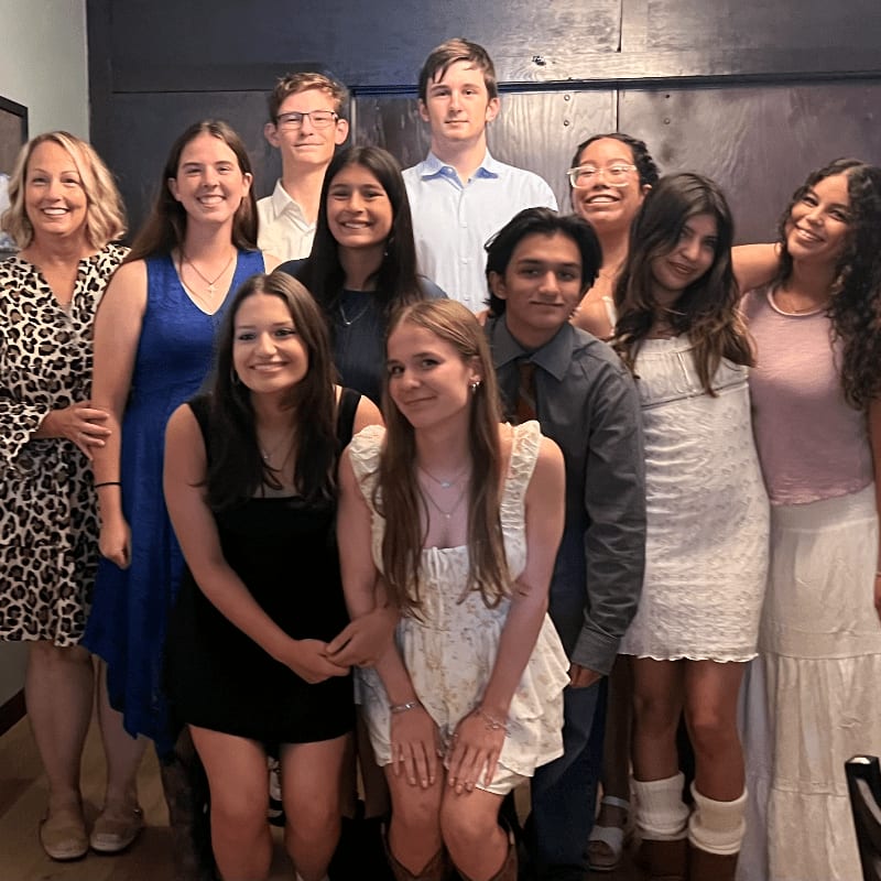 A group of smiling people, including adults and teenagers, pose for a photo in front of a dark, textured wall.
