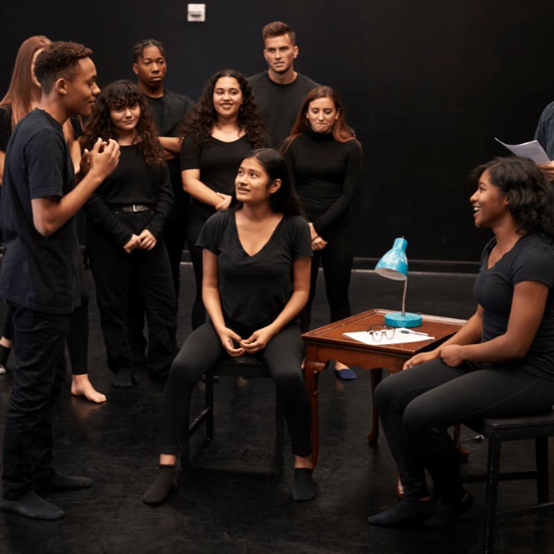 A group of young actors in black clothing rehearse a scene on a dark stage, with two individuals in the foreground engaged in dialogue near a small table.