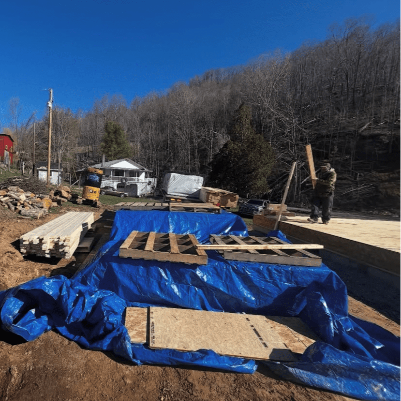 A construction site with lumber and building materials covered by a blue tarp in the foreground, and a house and wooded hillside in the background.