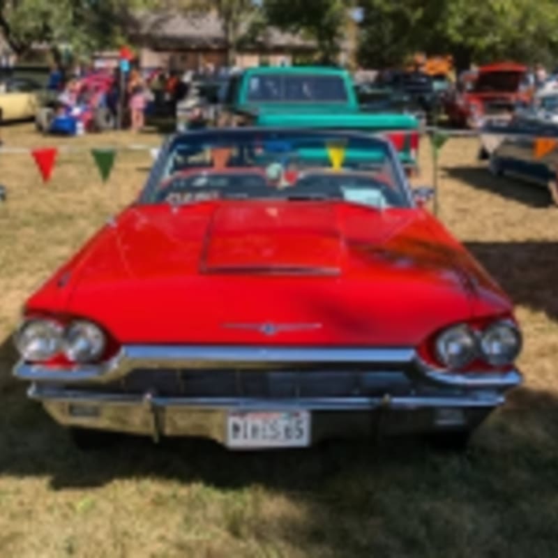 A bright red vintage convertible is parked on a grassy field with other classic cars and people in the background.