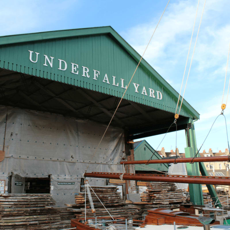 In the foreground, wooden planks are stacked near a boat, while a large green building with "UNDERFALL YARD" written on it stands in the background under a blue sky.
