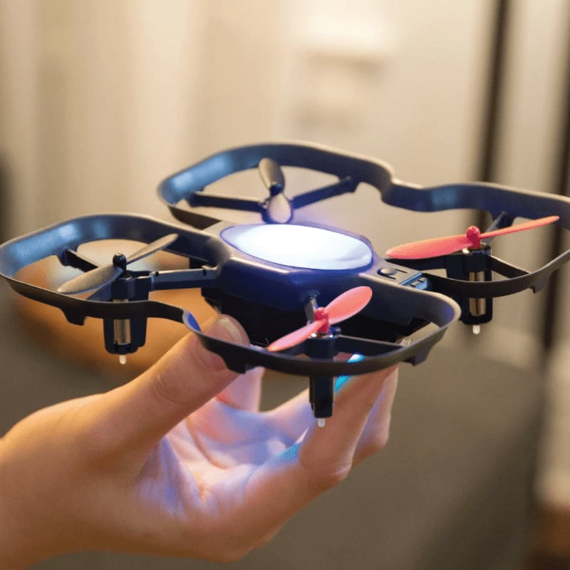A small drone with red propellers rests on a hand in the foreground, while a blurred, indoor background is visible behind it.