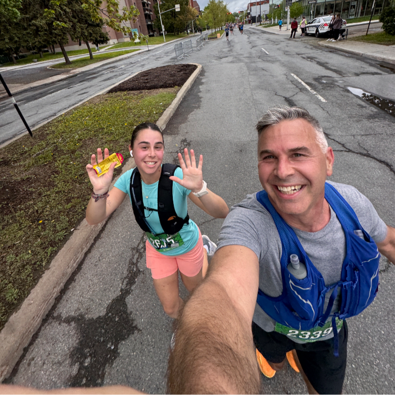 A man and a woman in running gear smile and wave while running down a street with buildings and trees in the background.