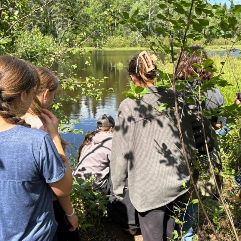 A group of people gather by a calm lake surrounded by lush green trees and foliage.