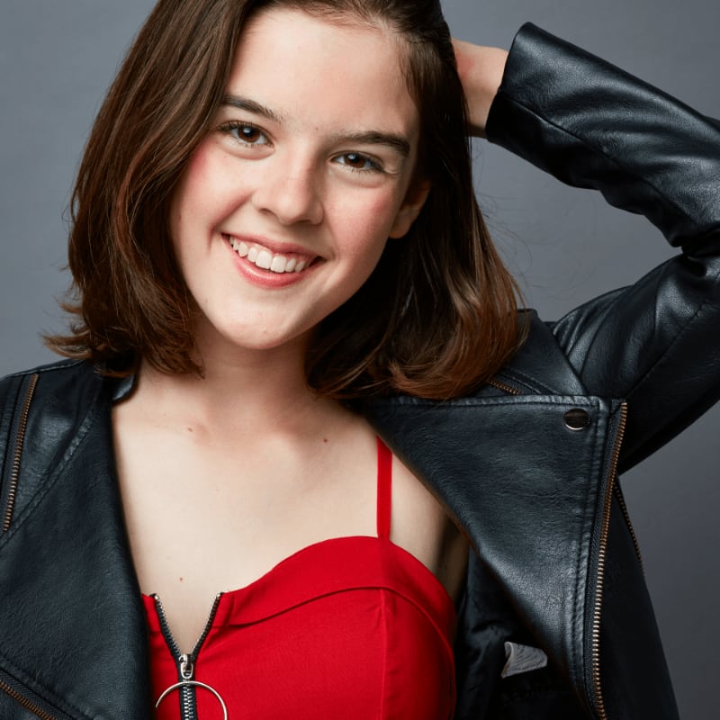 A young girl with brown hair smiles, wearing a red top and black leather jacket, against a plain gray background.