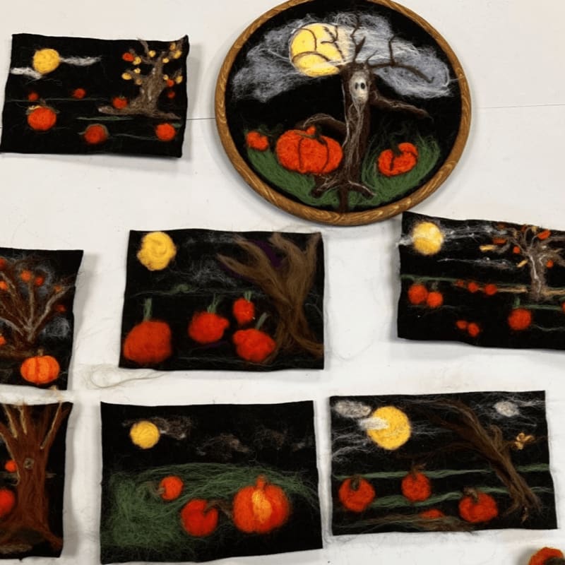 Felted artworks of pumpkins and spooky trees under a moonlit sky are displayed on a white surface.
