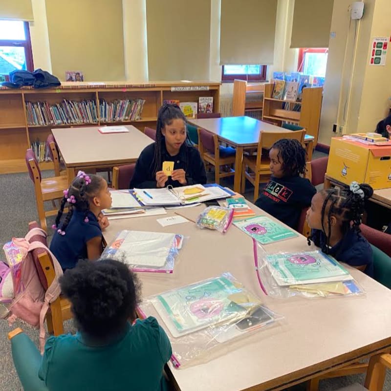A teacher holds up a flashcard to three young students sitting at a table in a library with bookshelves in the background.