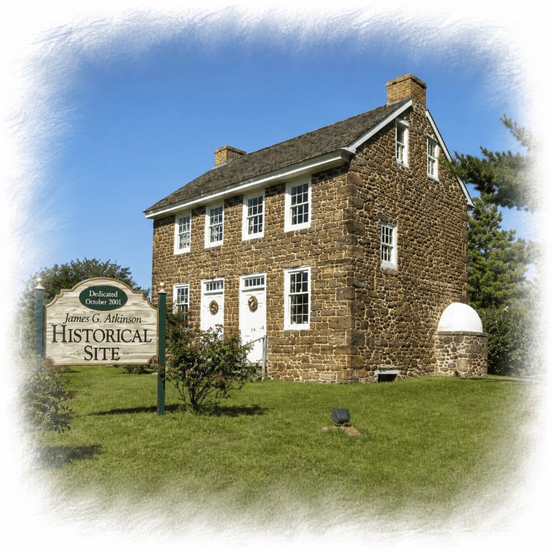 A historic stone house with white-trimmed windows and doors stands on a grassy lawn in front of a clear blue sky, with a wooden sign indicating it is a historical site.