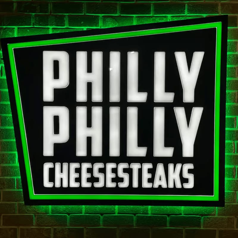 A neon-outlined sign with "PHILLY PHILLY CHEESESTEAKS" in white block letters is mounted on a brick wall.