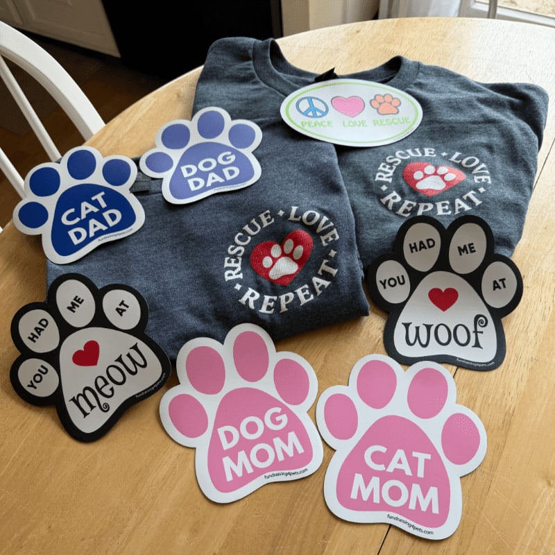 Paw print magnets with pet-themed slogans are scattered on a wooden table in front of a gray t-shirt with similar designs.