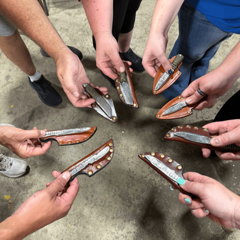 Eight hands hold up handmade knives with leather sheaths in a circle, with a concrete floor visible in the background.