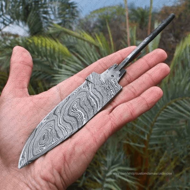 A hand holds a Damascus steel knife blade with a swirling pattern, set against a blurred background of palm fronds.