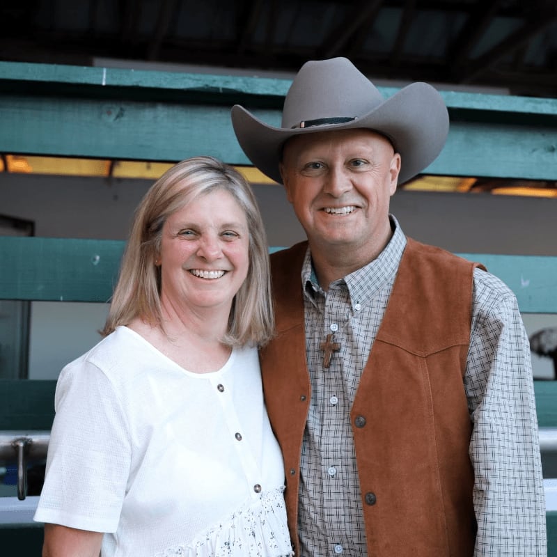 A smiling couple, a woman in a white top and a man in a cowboy hat and vest, stand in front of a green wooden structure.