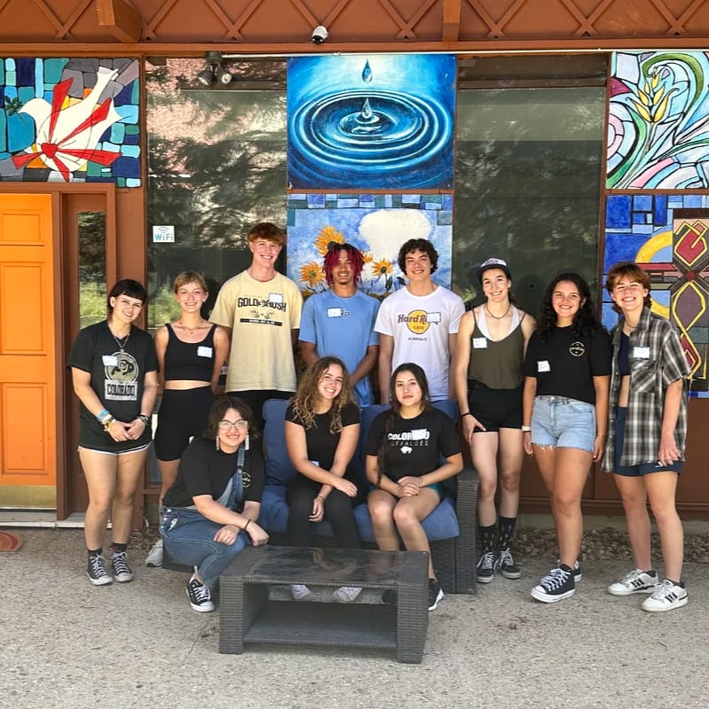 A group of young people pose for a photo in front of a colorful mosaic wall.