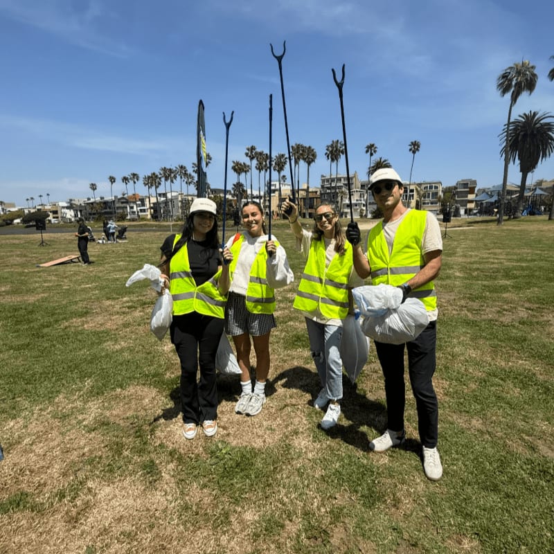 Four people in bright yellow vests stand on a grassy field holding trash grabbers and bags, with palm trees and buildings in the background under a clear blue sky.