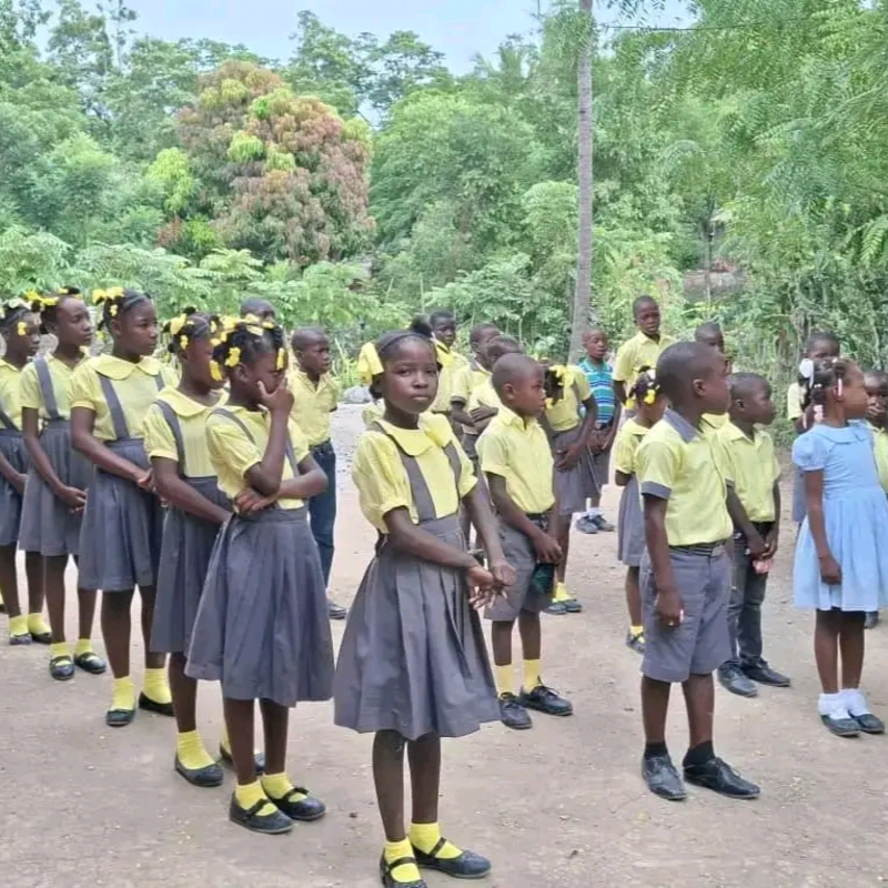 Children in yellow shirts and grey skirts stand in a line on a dirt path, with lush green trees and foliage in the background.